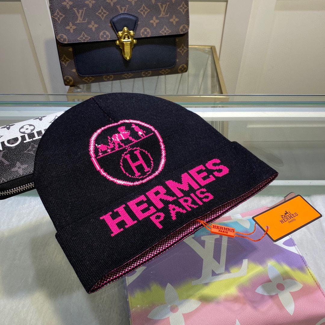 Black beanie with pink 'Hermes Paris' logo on a display table with a Louis Vuitton bag in the background.