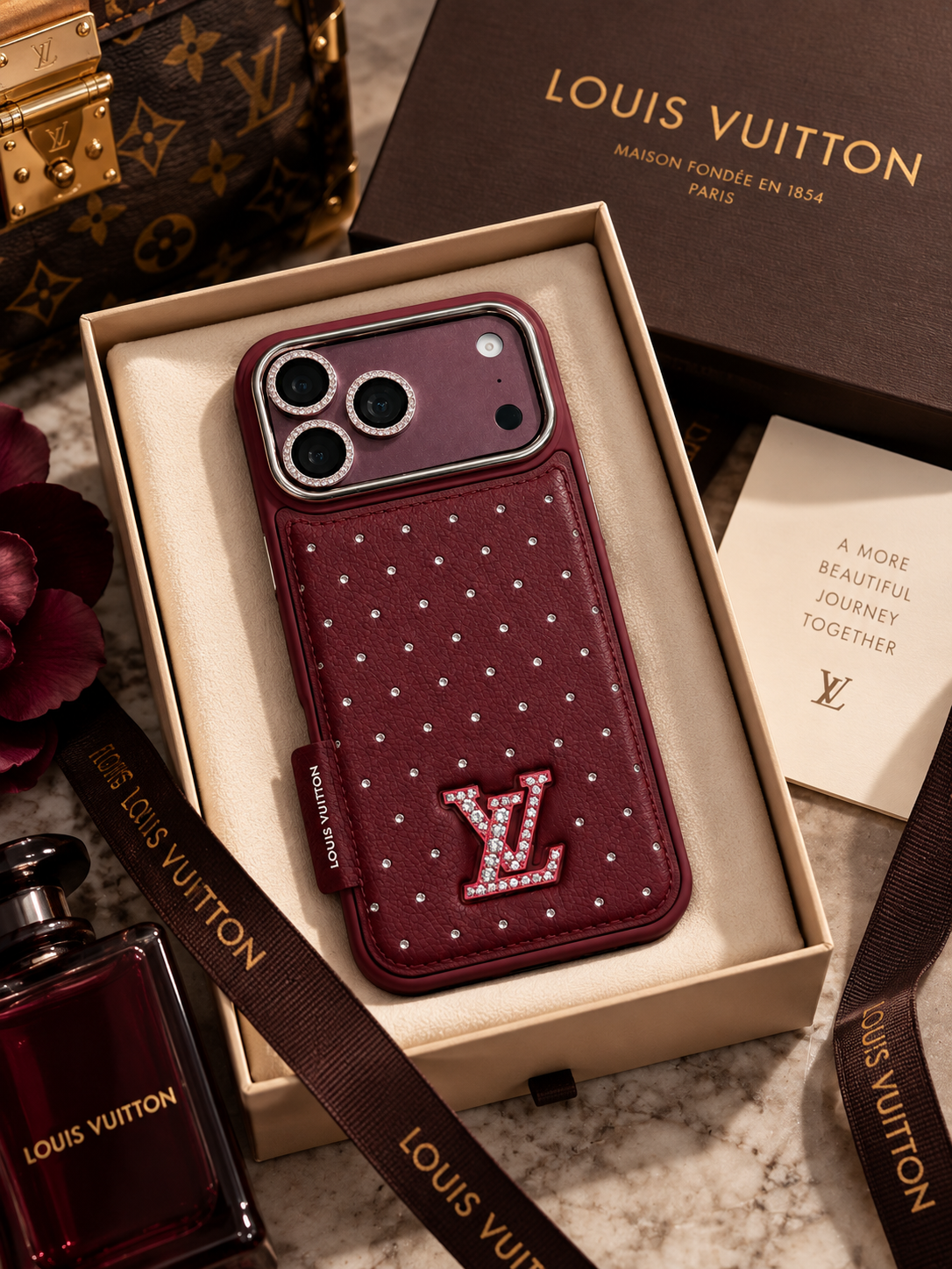 A Louis Vuitton leather phone case in deep burgundy with rhinestone dot details and a rhinestone-embellished LV logo, displayed inside an open beige presentation box. The box is surrounded by branded packaging, a Louis Vuitton perfume bottle, and a monogram bag on a marble surface.